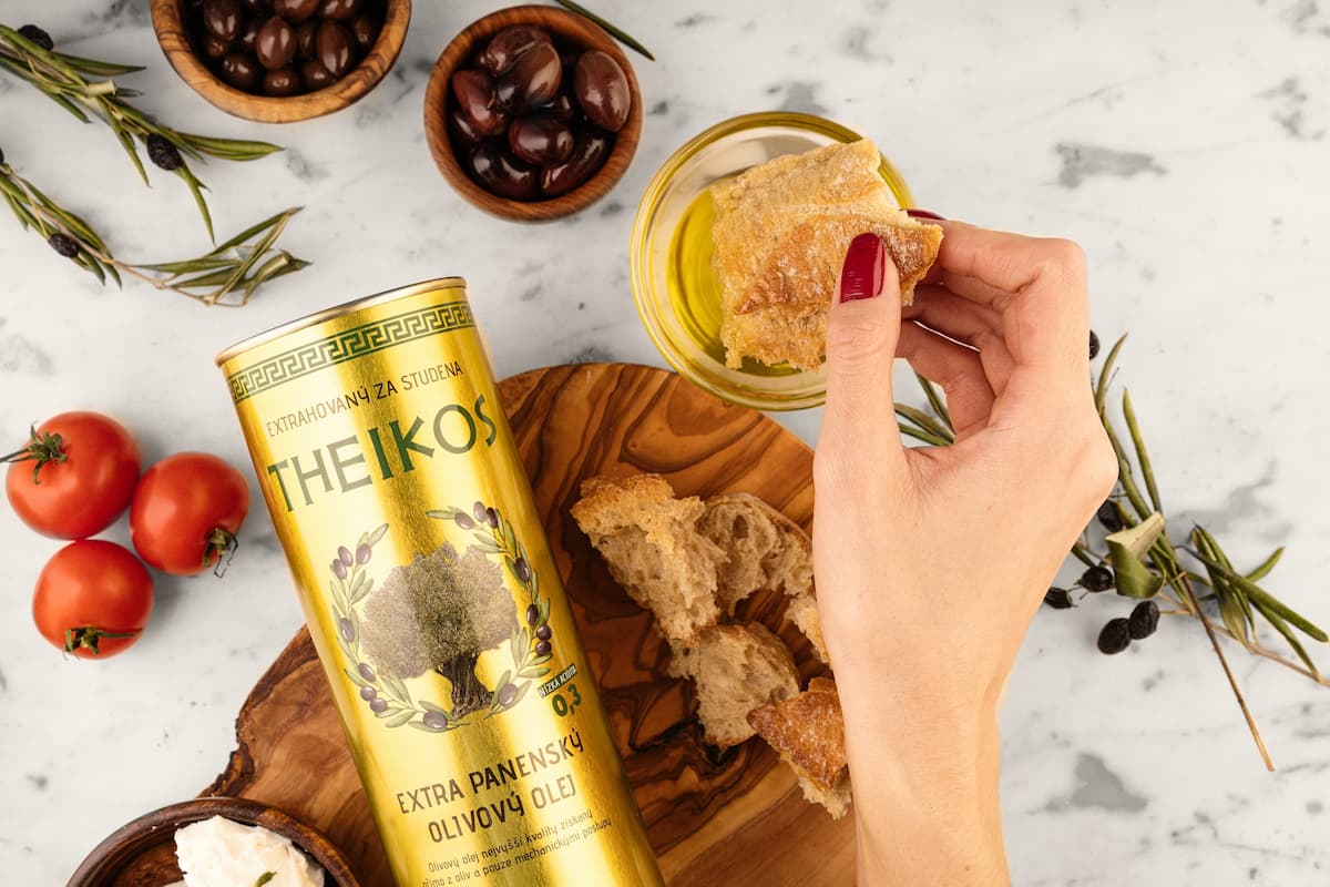 Ruka namáčí pečivo do extra panenského olivového oleje Theikos na dřevěném prkénku s olivami a rajčaty